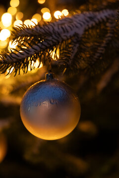 Golden ornament hanging on a tree branch with bright lights in the background during holiday season