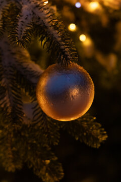 Christmas ornament hanging on a snow-covered tree branch at night with lights glowing in the background