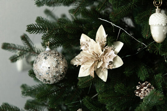 Decorations hang on a green Christmas tree with silver and white ornaments and flowers in a room