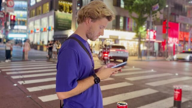 Young Man Standing at Pedestrian Crossing Using Smartphone at Night &ndash; Waiting for Traffic Light with City Lights Background