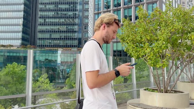 Young Blond Man Standing on Bridge with Smartphone and Skyscraper Skyline in Background &ndash; Summer Day in Modern City