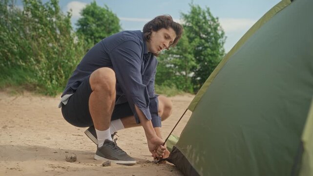 Caucasian young adult man squatting on sandy beach and securing peg of green camping tent. Footage is ideal for outdoor gear commercials and adventure travel marketing campaigns