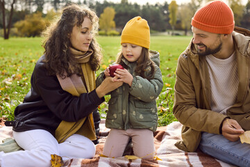 Fototapeta premium Happy family in autumn park, smiling daughter. Parents share an apple with their child on a picnic blanket. Warm colors and scarves show love and bonding. Family togetherness and seasonal joy.