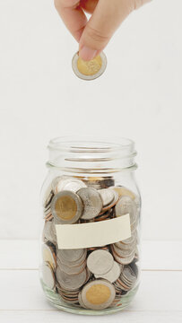 Woman's hand dropping ten Thai Baht coin into a glass jar full of coins. Business and finance concept. Vertical Photography.