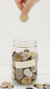 Woman's hand dropping ten Thai Baht coin into a glass jar full of coins. Business and finance concept. Vertical Photography.