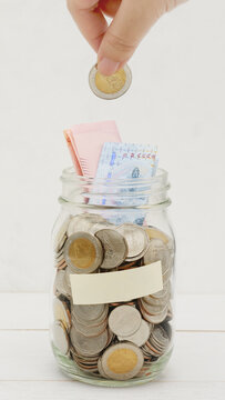 Woman's hand dropping ten Thai Baht coin into a glass jar full of coins. Business and finance concept. Vertical Photography.