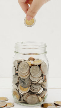 Woman's hand dropping ten Thai Baht coin into a glass jar full of coins. Business and finance concept. Vertical Photography.