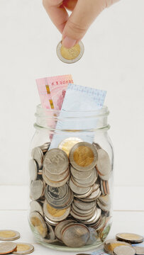 Woman's hand dropping ten Thai Baht coin into a glass jar full of coins. Business and finance concept. Vertical Photography.