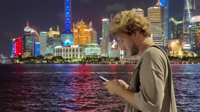 Summer Night at The Bund &ndash; Young Blond Man Standing on Waterfront with Smartphone, Reading, Scrolling and Taking Photos Against Skyscraper Skyline in Shanghai