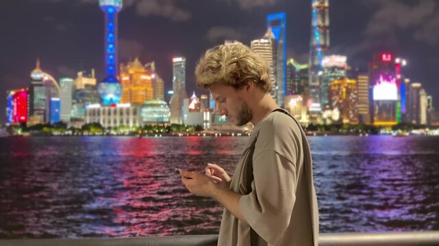 Summer Night at The Bund &ndash; Young Blond Man Standing on Waterfront with Smartphone, Reading, Scrolling and Taking Photos Against Skyscraper Skyline in Shanghai