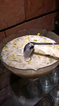 Vertical video of hand stirring refreshing Asian milky iced dessert with avocado and jelly in a large bowl at a catering buffet