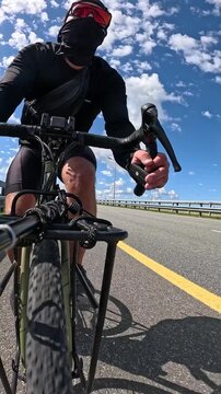 High speed vertical POV footage from a low angle of a focused male cyclist in a khaki t-shirt and backward baseball cap sprinting fast on the lower drop bars of a gravel bike along a highway.