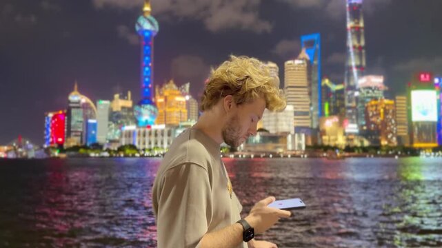Summer Night at The Bund &ndash; Young Blond Man Standing on Waterfront with Smartphone, Reading, Scrolling and Taking Photos Against Skyscraper Skyline in Shanghai