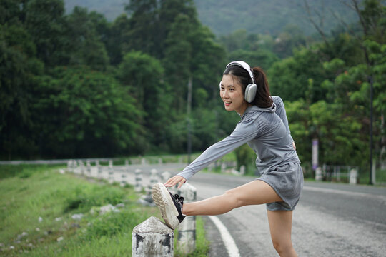 A medium shot of a cheerful Asian woman stretching her hamstring on a roadside post with green nature and mountains in the background.