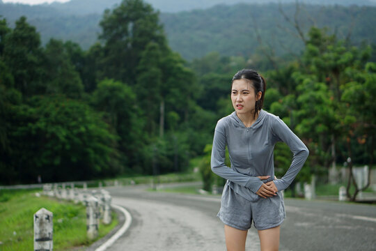 An Asian female runner bending forward while clutching her painful side stitch on a road near a green forest background.