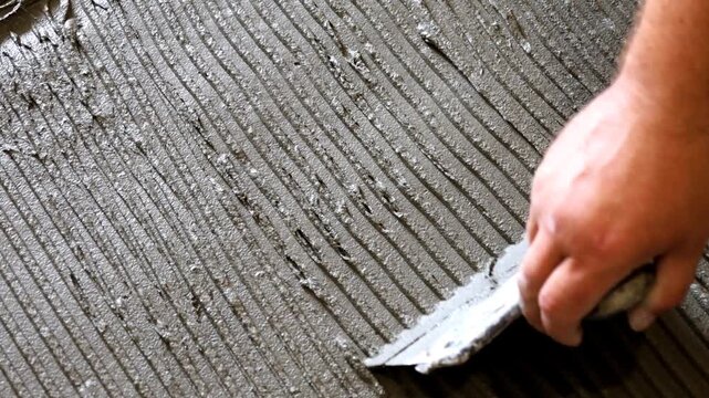 Close-up professional stock video showing a worker's hand carefully applying tile adhesive onto a floor using a notched trowel. The process of creating a ridged surface for subsequent tiling is visibl