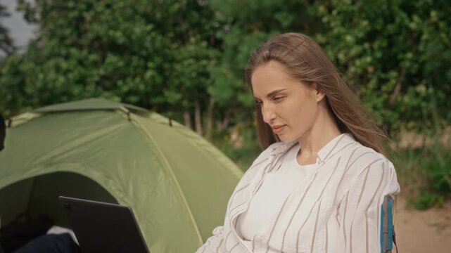 Caucasian young adult woman sitting in folding chair near camping tent, typing on laptop with camera tilting up. Clip is perfect for promoting remote work and digital nomad lifestyle