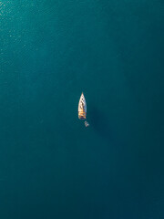 Bird's eye aerial view of a lone sailboat with a small dinghy floating on calm deep blue water, surrounded by expansive copy space. © Krisztin