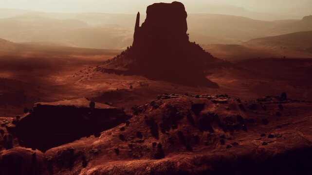A view of Monument Valley during sunset shows tall rock formations in the distance. The landscape is vast with warm tones and shadows across the ground.