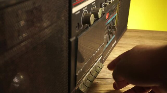 A close side angle reveals a male hand removing a clear audio cassette from a dusty vintage black radio boombox sitting on a wooden desk near a bright yellow wall.