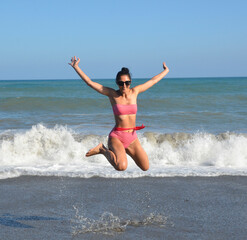 Fototapeta premium A woman enjoying a playful moment beside the sea, jumping over wet sand as waves reach the shore, perfect for vacation and summer lifestyle concepts.
