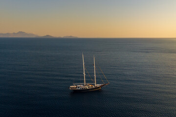 Fototapeta premium Aerial view of a two masted sailing yacht on calm open water, illuminated by warm golden hour light with distant mountainous islands along the horizon.