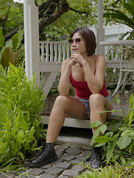 Thoughtful Asian woman in red tank top relaxing amidst green foliage