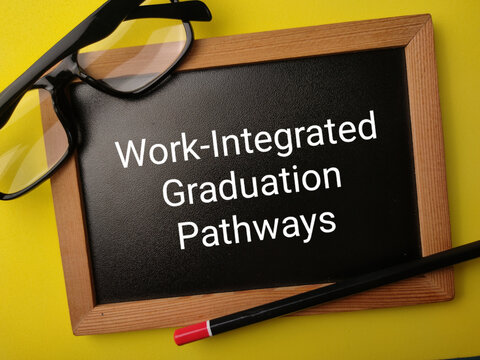 A wooden framed blackboard reading Work-Integrated Graduation Pathways lies with glasses and pencil on yellow