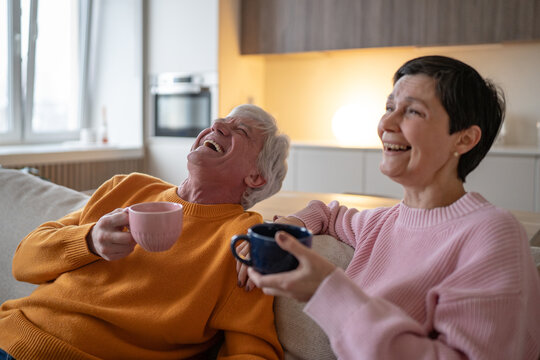 Happy senior couple at home together sitting on couch relaxing enjoying watching funny movie on tv, cheerful positive elderly husband and wife laughing at scene while resting on couch with drinks.