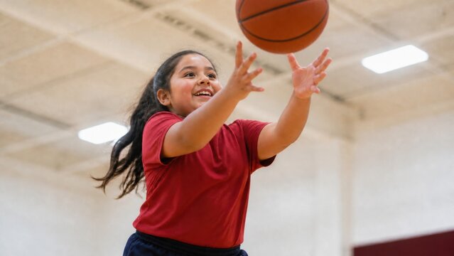 Happy girl catching basketball during indoor sports practice. Active childhood, athletic development, teamwork, healthy lifestyle, and youth recreation concept.