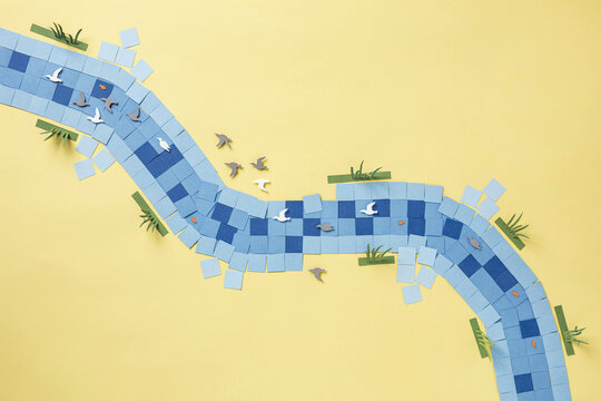 Top Down View of a Stylized Paper Craft River Scene with Birds and Fish Created from Geometric Blue Squares on a Textured Yellow Background