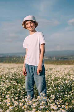Smiling teen boy wearing bucket hat standing in field of Matricaria chamomilla flowers during sunny spring day