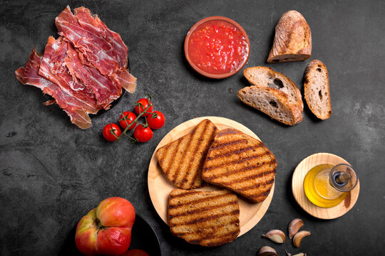 Toasted bread slices with cured Spanish ham, fresh tomatoes, and grated tomato sauce on dark surface, traditional healthy Mediterranean breakfast