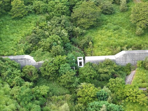 Ancient stone wall cuts through lush green forest. Trees frame the path with dense, vibrant foliage. Narrow, arched opening reveals history's quiet passage. Wind rustles leaves around ancient