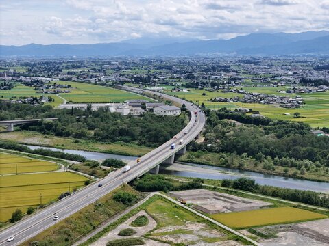 Aerial view of a highway crossing a river. Vehicles flow steadily over bridges. Fertile fields flank the waterway. Distant mountains frame the horizon. Urban sprawl blends with green landscapes