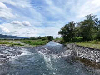 Clear river rushes over smooth stones under bright sky. Clouds drift lazily across blue horizon above green hills. Riverbank grows wild with tall grass and leafy trees © Masakazu Tokashiki