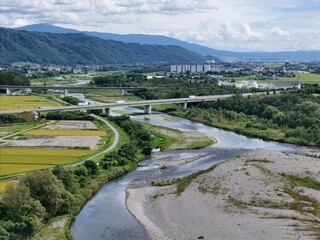 River winds through green fields, bordered by distant hills. Clouds drift above, casting gentle light on the landscape. Bridge spans the water, connecting farmland and town © Masakazu Tokashiki