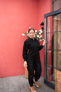 Stylish woman in black jumpsuit enters glass doorway beside blooming white roses against pink wall