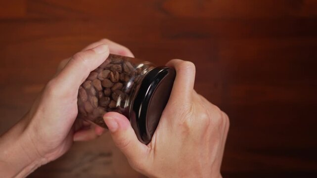 Centered top view of woman hands holding coffee bean glass jar, straining wrist to twist tight cap until seal loosens, setting jar down on wooden desk, breakthrough concept.⁠