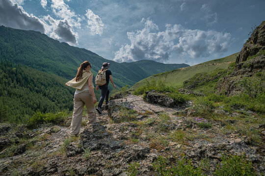 Rear view of two young women with backpacks hiking on a rocky mountain path overlooking a lush green valley under a cloudy sky, female friendship and active travel concept