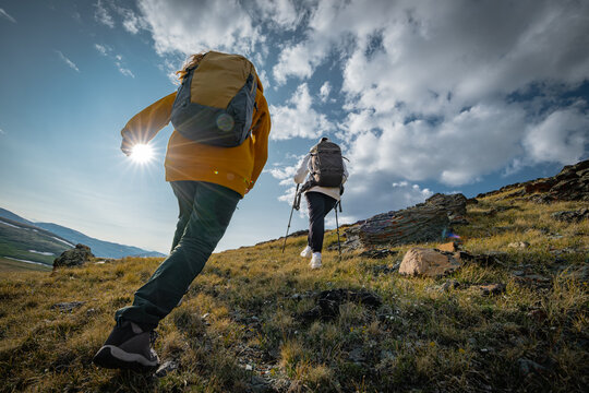 Rear view of two young backpackers with hiking gear walking up a steep grassy mountain hill under a blue cloudy sky with sun flare, active travel and exploration concept