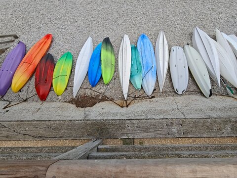 A top-down view of vibrant multicolored kayaks and canoes neatly lined up side by side on a pebble beach, showcasing bright shades of red, orange, green, blue, and white for water sport activity.