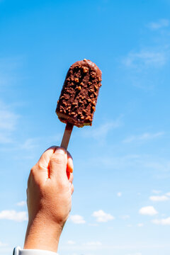 Hand holding crisp chocolate ice cream bar with nuts on wooden stick against clear blue sky in summer. Delicious cold sweet dessert snack outdoors on bright sunny day. Vertical