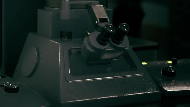 A microscope sits on a lab table in a science lab. Researchers are using it to look at samples. The lighting in the lab is dim. Tools and equipment surround the microscope.