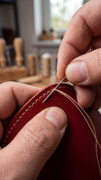 Close-up of male hands sewing dark red leather with a needle and thread in a workshop. Craftsmanship, handmade leather goods process, professional tool detail concept.