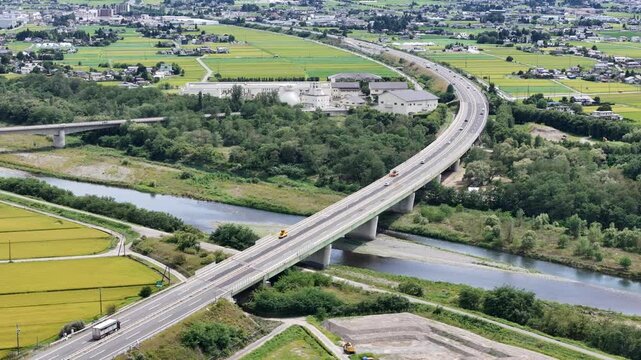 Aerial view of highway crossing a river. Green fields flank the road and bridge. Vehicles move steadily on the curved lanes. River flows calmly beneath the concrete structure