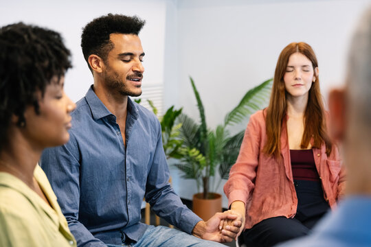 Group of people sitting in a circle, holding hands, and meditating with closed eyes, finding emotional support and community in a therapy session for improved mental health and wellness