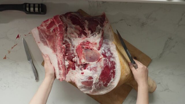 Hands guiding knife to portion bone-in pork chunk on cutting board during home meat prep