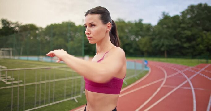 Young athletic woman in sportswear warming up and doing exercises on the stadium running track in the evening at sunset