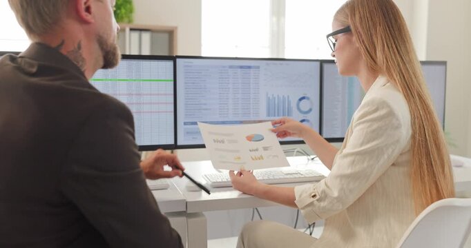 Colleagues businesswoman manager conduct analysis of report. They review charts on dual monitors, discussing finance metrics and budgeting choices. Clear teamwork and finance analysis concept.